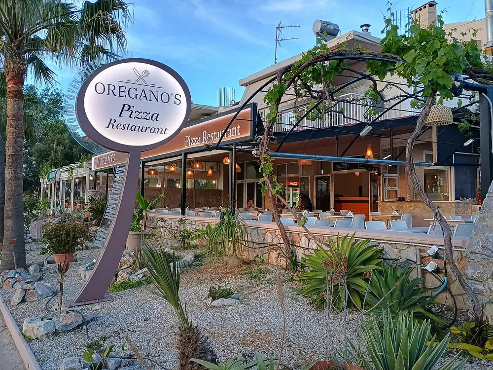 Oregano’s Pizza Restaurant entrance and outdoor seating area surrounded by palm trees and local greenery in Agii Apostoli, Chania.